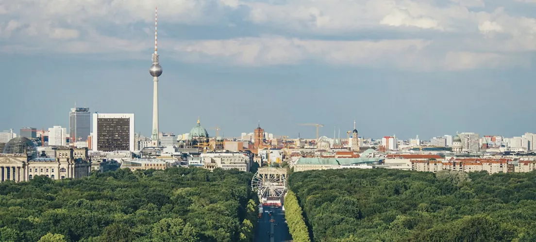 Panoramic view of the Berlin skyline with the Fernsehturm TV tower rising above the Tiergarten under a partly cloudy sky.